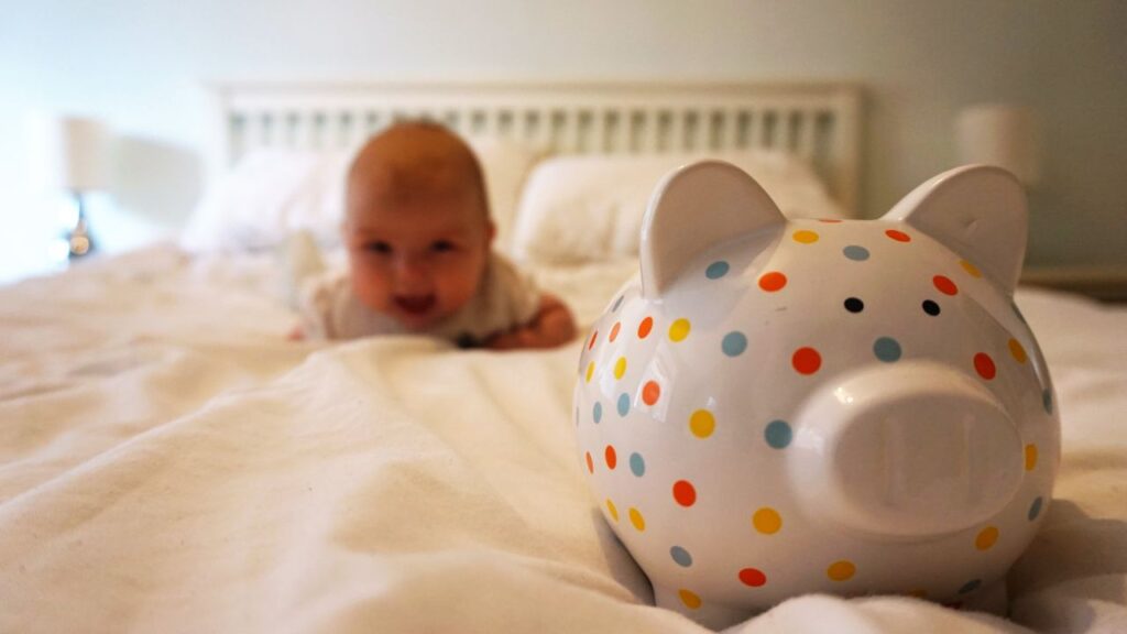 Baby liegt auf einem Bett und krabbelt auf ein Sparschwein zu